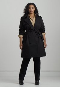 Svart trench coat med dubbelknäppt design, knappdetaljer och bälte i midjan. Bärs över en beige skjorta, matchat med svarta byxor och stövlar.