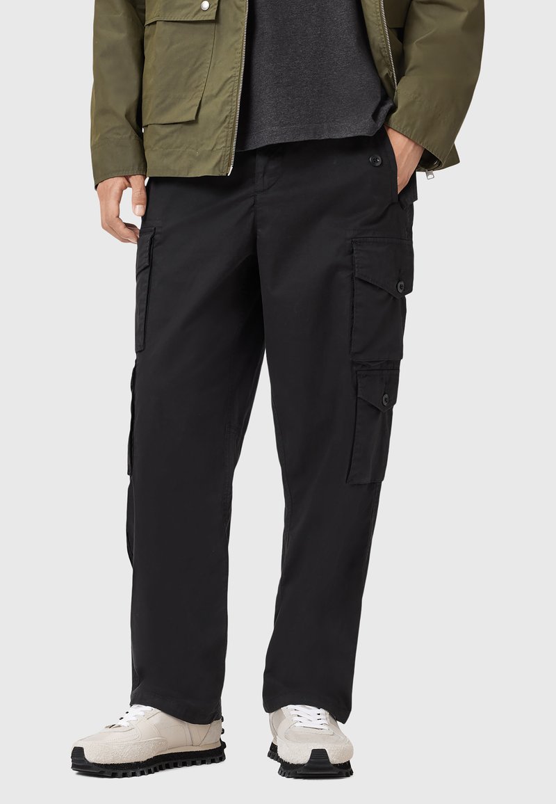 Pantalons cargo noirs avec plusieurs poches latérales, coupe ample et texture lisse, associés à des baskets beiges et noires pour le contraste.
