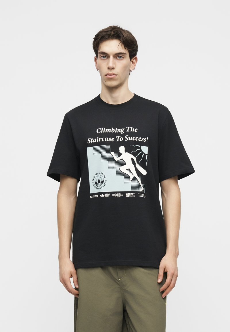 Junger Mann trägt schwarzes T-Shirt mit Grafik einer Person, die eine Treppe hinaufsteigt, und dem Text "Die Treppe zum Erfolg erklimmen!"