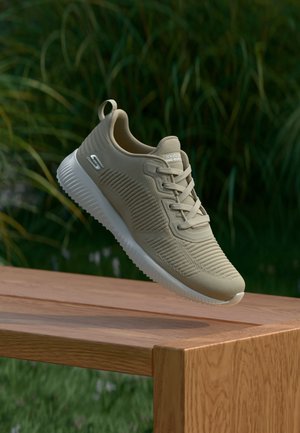 Beige Skechers sneaker with ribbed texture and white sole floating above wooden bench outdoors with green plants in background.