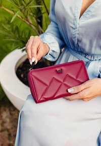 Fuchsiová kožená peněženka s geometrickým patchworkovým designem, stříbrnými doplňky a zipovým uzávěrem. Ruka držící peněženku proti světlému textilnímu pozadí.