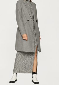 Grauer zweireihiger Mantel mit taillierter Passform, über einem grauen Strickkleid mit einem seitlichen Schlitz getragen. Weiße Knöchelstiefel runden das Outfit ab.