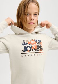 Svetlo bež pulover s kapuco in grafiko spredaj, z velikim napisom "JACK & JONES WORLDWIDE" v različnih barvah. Mehka tkanina, oprijeta oblika.