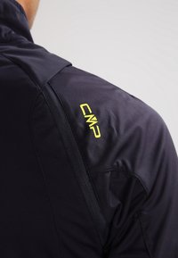 Marineblå jakke med en blød, glat overflade. Har en lynlåslomme og et gult "CMP" logo på skulderen for synlighed og branding.