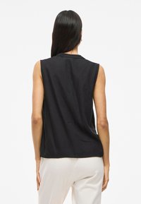 Musta ilmanhihaista toppi, joka on valmistettu sileästä kankaasta ja jossa on rento istuvuus. Siinä on pyöreä kaula-aukko ja hieman pidempi takahelma.