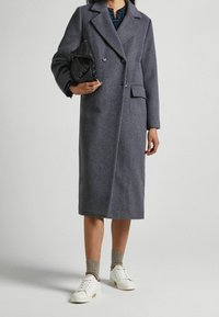 Abrigo de lana gris con diseño de doble botonadura, solapas muescas, bolsillo lateral y corte hasta la rodilla, combinado con zapatillas blancas y un bolso de mano negro.