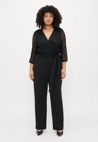 Svart jumpsuit med en strukturerad spetsomslag vid bysten, trekvartslånga ärmar och bälte i midjan; kombinerad med svarta klackar med spetsig tå.