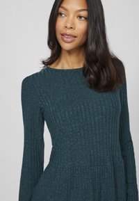 Türkises, geripptes Langarmkleid mit tailliertem Oberteil und schwingendem Rock, ausgestattet mit einem runden Ausschnitt und strukturierten Stoffdetails.