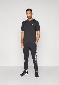 Juodas trumpos rankovės marškinėliai su baltu logotipu ant krūtinės, derinami su juodomis sportinėmis kelnėmis su baltais šonų dryžiais ir juodais sportiniais batais.