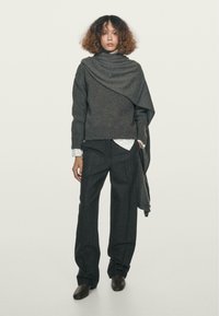 Grauer Strickpullover mit asymmetrischem Drapieren, über einem weißen Hemd mit Kragen getragen. Dunkelgraue, weite Hose und dunkle Schuhe. Schlicher Hintergrund.