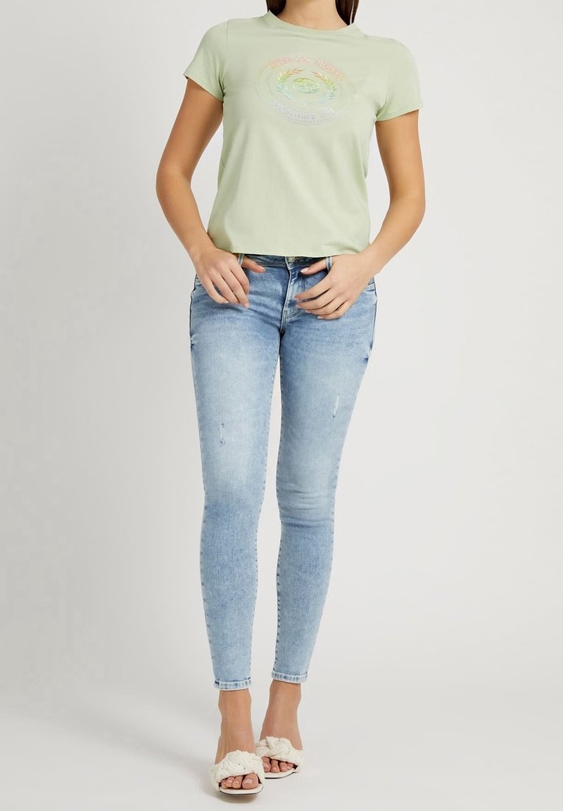 T-shirt vert clair à manches courtes avec un graphique circulaire multicolore, associé à un jean skinny bleu clair avec des accents déchirés et des sandales blanches à enfiler.