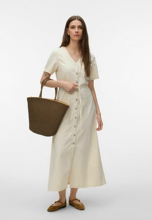 Naine kandes kreemjat keskpikkade nööpidega kleiti, hoides suurt pruuni ja musta triibulist kootud käekotti, jalas pruunid tasased kingad.