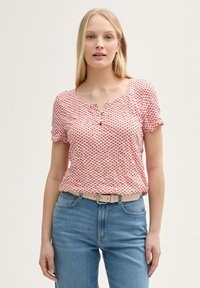 Haut à manches courtes rouge et blanc à motifs avec un col rond et un détail de bouton, associé à un jean en denim bleu clair et une ceinture beige.