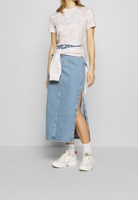 Světle šedé tričko s tie-dye vzorem, džínová maxi sukně s bočními rozparky a bílé tenisky s červenými akcenty. Neformální, vrstvený outfit s volným střihem.