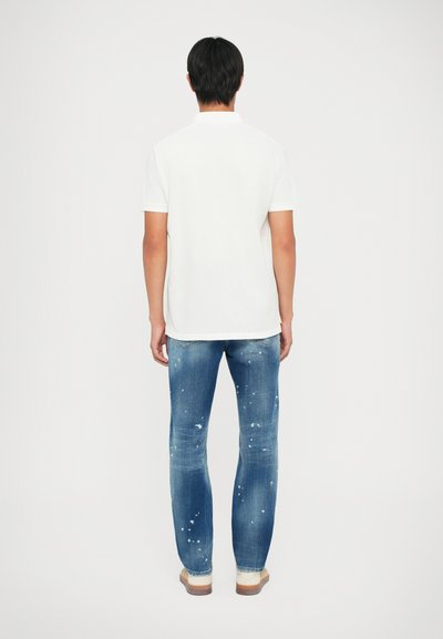 Fehér rövid ujjú polo póló, kék, kopott denim farmerrel, amelyen festékfröccsenések találhatók, és egyenes szabású. Lábbeli látható.