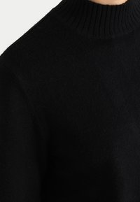 Svart stickad tröja med en hög ribbad krage. Tyget verkar mjukt med en något strukturerad yta, vilket betonar en minimalistisk design.