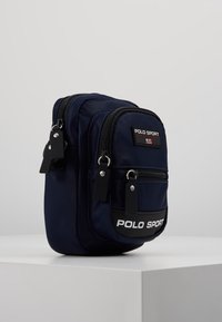 Zaino in nylon navy con due scomparti con cerniera, patch con logo frontale, accenti neri e tessuto texturizzato. Design rettangolare compatto.