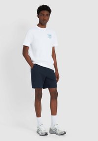Witte katoenen T-shirt met blauwe grafische details, gecombineerd met marineblauwe textuur shorts. G gedragen met witte sokken en grijze sneakers.