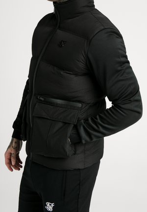 Sort isoleret vest med høj krave, quiltet tekstur, lynlåslukning foran og en klaplomme på siden. Bæres over en langærmet skjorte.