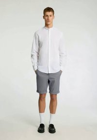 Λευκό πουκάμισο με μακριά μανίκια και κουμπωτό γιακά, συνδυασμένο με γκρι σορτς. Το μοντέλο φορά λευκές κάλτσες και μαύρα παπούτσια. Απλή φόντο.