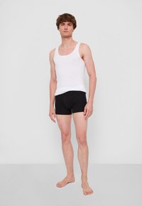 Vit bomullstopp utan ärmar och svarta boxershorts på en modell. Har en rund halsringning och en figurnära siluett. Barfota, stående mot en enfärgad bakgrund.