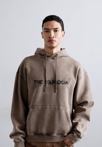 Brauner Hoodie aus weichem Stoff, ausgestattet mit einer Fronttasche, Kordelzügen und einem grafischen Druck mit verschiedenen Schriftarten auf der Brust.