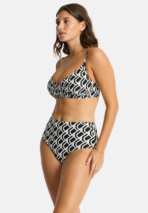 Kobieta stoi bokiem w czarno-białym bikini z geometrycznym wzorem i majtkami z wysokim stanem na jednolitym tle.