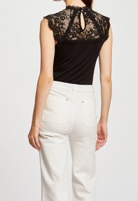 Mujer vestida con una blusa sin mangas negra con detalles de encaje floral en la parte posterior y pantalones blancos de cintura alta, de pie contra un fondo sencillo.