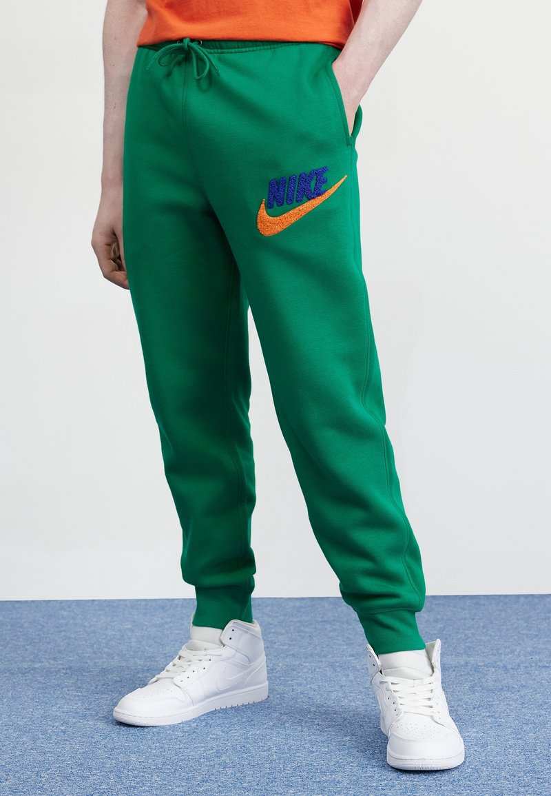 Zelené tepláky z měkké látky, s modrým logem „NIKE“ a oranžovým znakem swoosh. Mají elastický pas a kapsy.