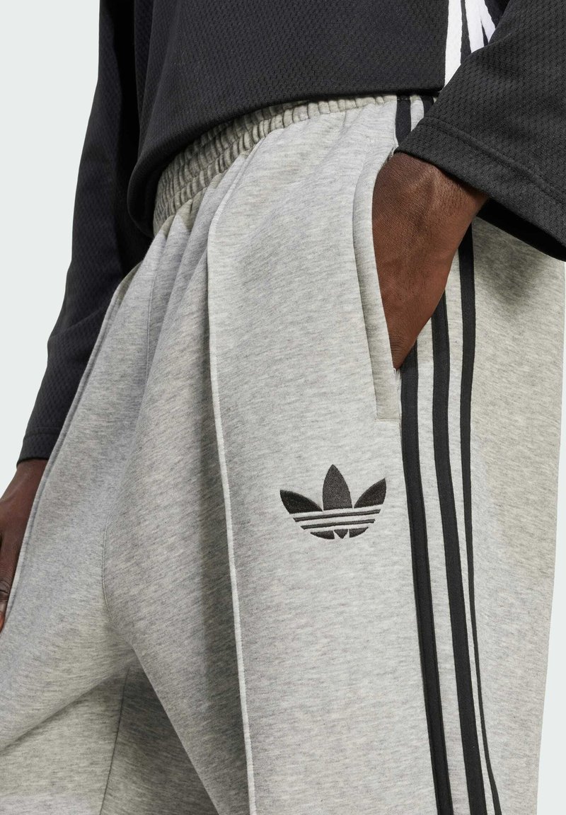 Pantalones de chándal grises de tela suave, con un logo negro de Adidas, bolsillos laterales y acentos en franjas negras a lo largo de los lados.