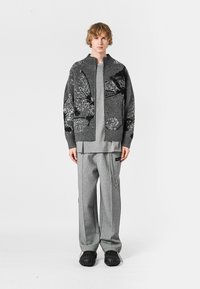 Cárdigan de punto gris con cierre de cremallera y patrones en blanco y negro, combinado con pantalones grises de gran tamaño, usado con zapatillas negras texturizadas.