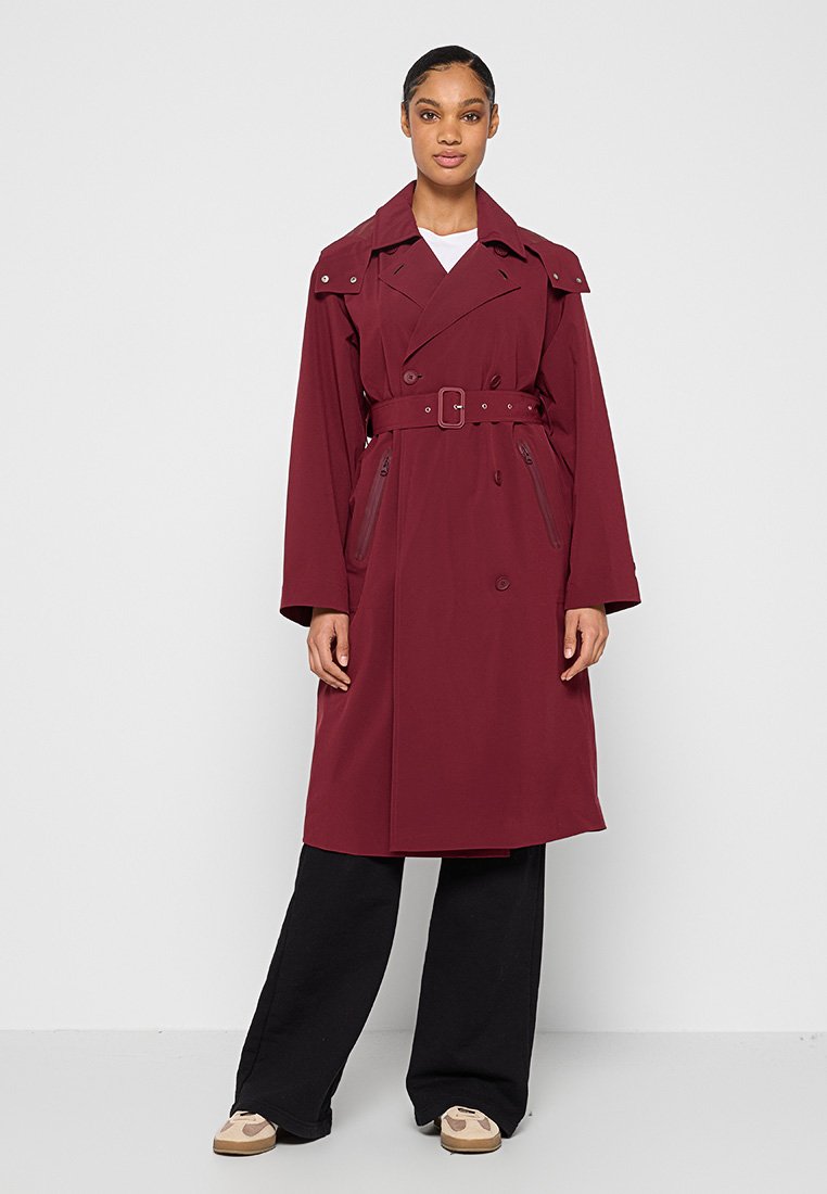 Vrouw in een lange bordeauxrode trenchcoat met riem, zwarte wijde pants en beige sneakers, staand tegen een effen lichte achtergrond.