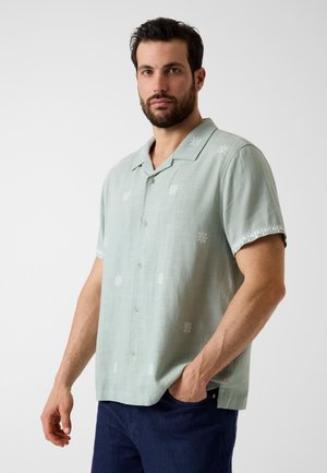 Mann mit dunklem Haar und Bart trägt ein hellgrünes kurzärmeliges Hemd mit weißen Blumenstickereien und dunkelblaue Hose, die Hand in der Tasche.