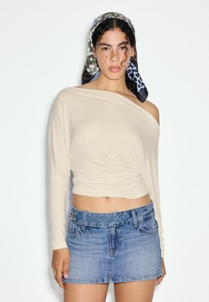 Femme aux cheveux bouclés portant un foulard à motifs, un haut crème épaules dénudées et une mini-jupe en jean bleu, debout devant un fond clair uni.