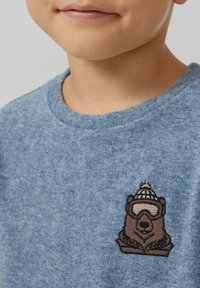 Ein Kind trägt einen blauen Rundhals-Pullover mit einem gestickten braunen Bärenaufnäher, der eine Skibrille und eine Wintermütze auf der Brust trägt.