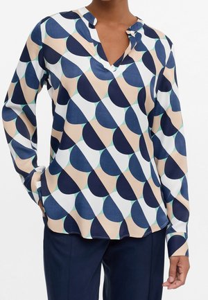 Blouse met lange mouwen, V-hals en geometrisch patroon van marineblauw, beige, teal en witte halve cirkels, gedragen met donker marineblauwe broek.