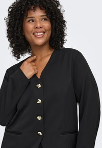 Čierny blejzer s výstrihom do tvaru V, zlatými dekoratívnymi gombíkmi a jemnými bočnými vreckami. Jemná látka s priliehavým strihom.