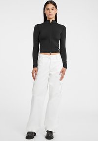 Top corto nero con zip e maniche lunghe abbinato a pantaloni cargo bianchi a gamba larga, dotati di tasche e stivali platform neri. Color solidi, texture liscia.