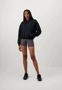 Svart bomullshoodie med framficka och snörning, i kombination med grå shorts. Modellen står i svarta sneakers mot en neutral bakgrund.