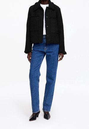 Person iført en sort tekstureret jakke, hvid skjorte, blå lige-benede jeans og sorte hælsko, stående mod en hvid baggrund.