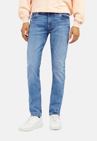 Jean en denim bleu clair usé avec une coupe slim, présentant des accents effilochés et un design traditionnel à cinq poches, associés à des baskets blanches.