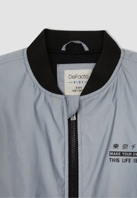 Svetle šedá bomber bunda s černým žebrovaným límcem, předním zipem a potištěným textem černou barvou. Materiál se zdá být hladký a mírně lesklý.