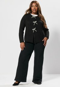 Svart stickad cardigan med tre texturerade krämfärgade rosetter framtill, matchad med vida svarta byxor. Enkelt, avslappnat utseende.