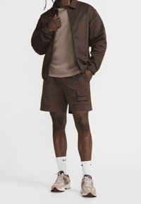 Brown athletic jacket with zip, matching brown t-shirt, cargo shorts with pockets, white socks, and beige sneakers. Minimalistic design.