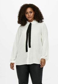 Blusa blanca de manga larga con un cuello clásico, detalle de pajarita negra en el cuello y una tela suave y lisa. Presenta un ajuste relajado.