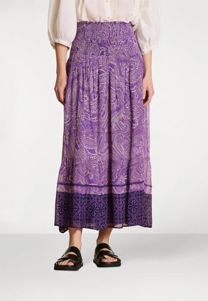 Maxi jupe à motif cachemire violet avec une taille smockée, présentant une bordure violet plus foncé à l'ourlet, associée à des sandales noires.
