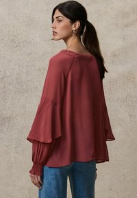 Rote, fließende Bluse mit Glockenärmeln, leichtem Stoff, gerafftem Ausschnitt und gepufften Manschetten, kombiniert mit blauen Jeans.