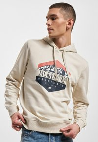 Cremefarbener Kapuzenpullover mit einem grafischen Druck, der Berge und den Text "JACK&JONES ORIGINALS" zeigt. Mit gerippten Bündchen und Saum, aus Baumwollmaterial.