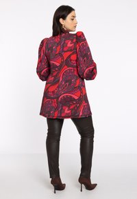 Rode en paarse tuniek met patroon en poffende mouwen, gecombineerd met zwarte leren broek en bruine haklaarzen. Glad stoftextuur.