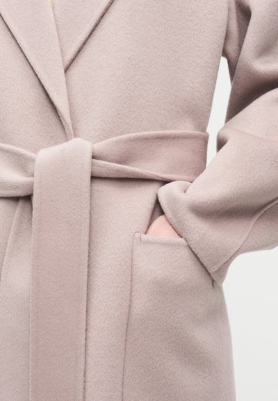 Κοντινό πλάνο ενός ανοιχτό ροζ μάλλινου παλτού με δεμένη ζώνη και ένα χέρι που ξεκουράζεται μέσα στην δεξιά τσέπη.